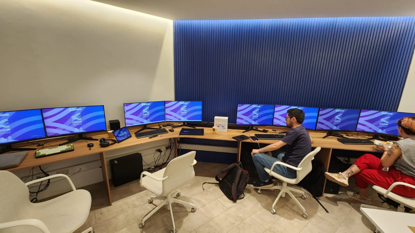 Sala CEGIR operator bay: two operators at a long wood desk with dual-monitor workstations running CEGIR dashboards.