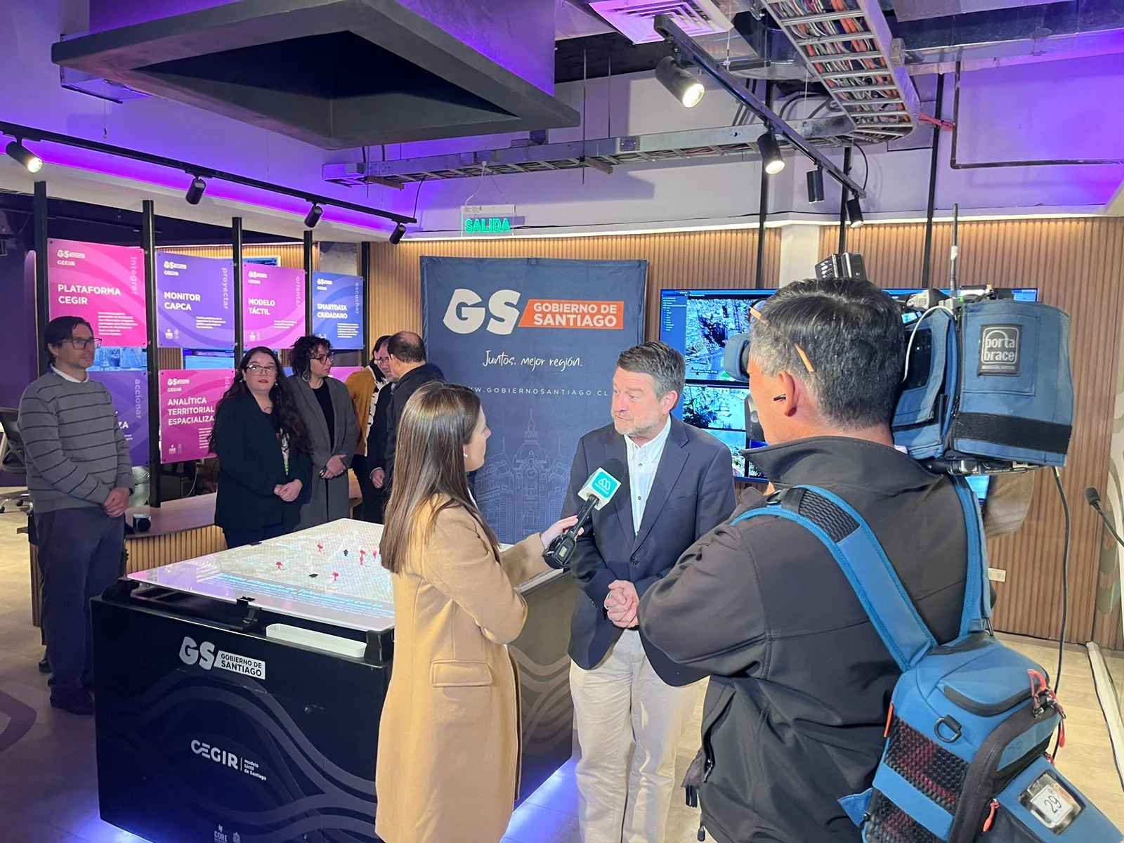 The Governor of Santiago giving a press interview inside the Sala CEGIR with backlit CEGIR product panels and the tactile table visible behind.