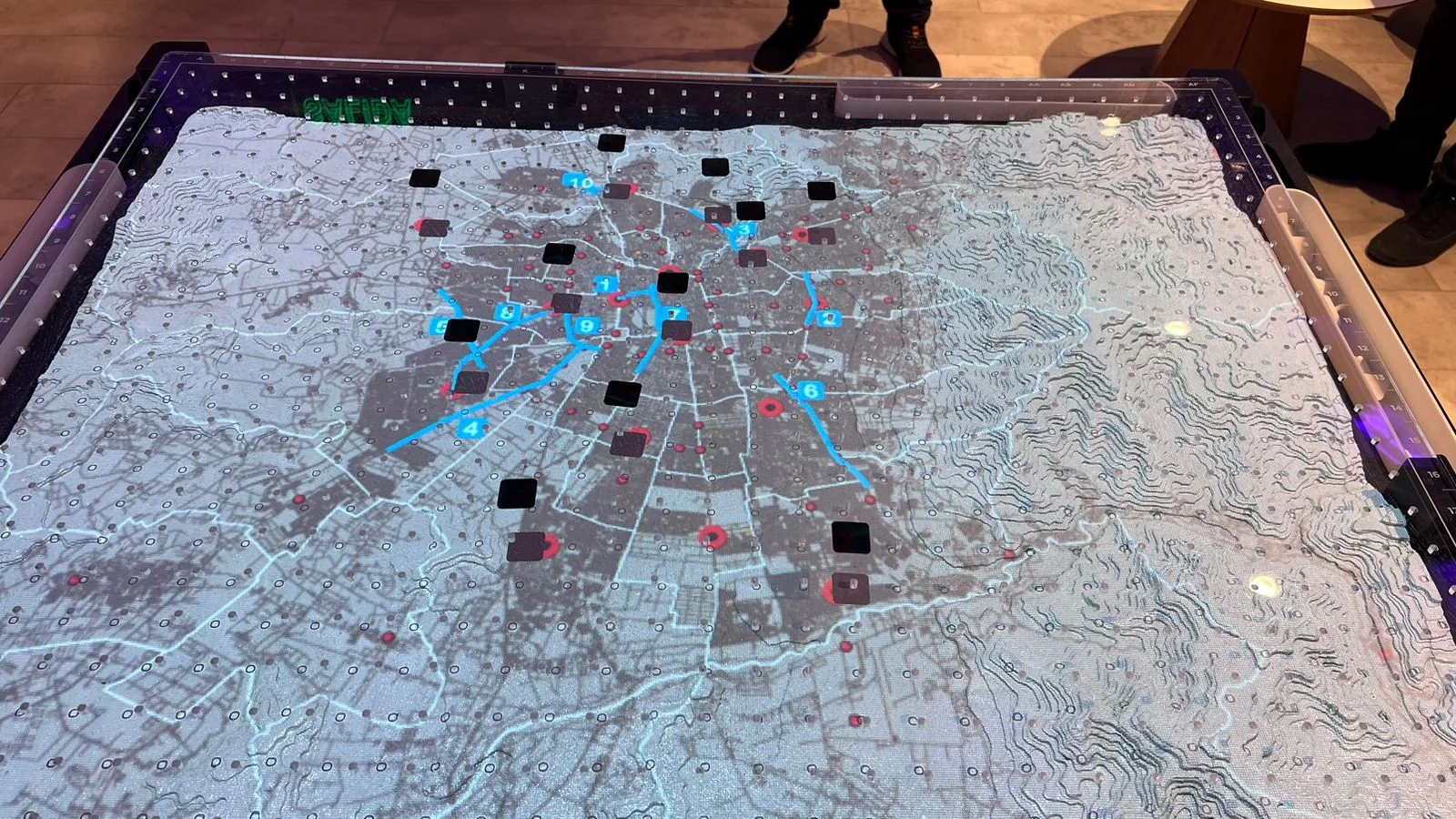 Tactile table projecting a Santiago street map with cyan flow-lines connecting tangible tokens placed at key nodes.